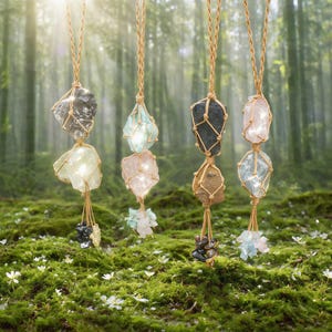 May include: A collection of gemstone pendants suspended from woven cords. Each pendant features a large, raw crystal paired with smaller, complementary stones. The crystals vary in color, including clear, pink, and smoky gray, set against a mossy forest backdrop.
