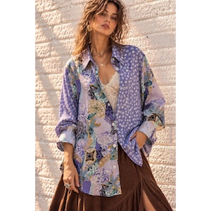 Può includere: Una camicia a maniche lunghe con bottoni in tonalità lavanda e viola, caratterizzata da un motivo patchwork misto di disegni floreali e geometrici. La camicia è indossata sopra una canotta in pizzo bianco e una gonna marrone scuro con spacco.