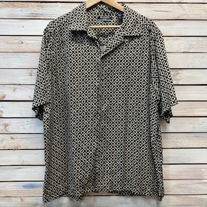 Puede incluir: Una camisa de manga corta con botones y un estampado geométrico en negro y beige. La camisa está colgada en una percha de madera. El nombre de la marca "BRANDINI" es visible en el cuello.