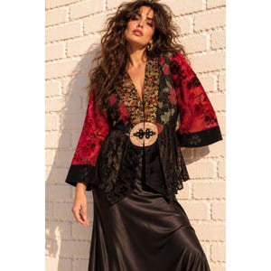 May include: A long-sleeved jacket featuring a mix of red and black floral patterns, lace panels, and a central beige circular closure with black knot detailing. The garment has a deep V-neckline and a flared silhouette, paired with a black satin skirt.