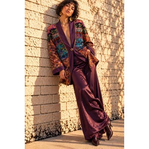 May include: A long-sleeved blazer in deep purple featuring a single-button closure and a vibrant, multi-colored patterned fabric on the sleeves and upper panels, paired with matching wide-leg satin trousers and dark brown pointed-toe boots.