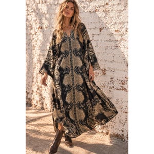 May include: A floor-length kaftan dress featuring a black and beige ornate paisley pattern, a deep V-neckline, and wide, flowing sleeves. The garment is paired with brown leather ankle boots, creating a cohesive look with a loose, draped silhouette.
