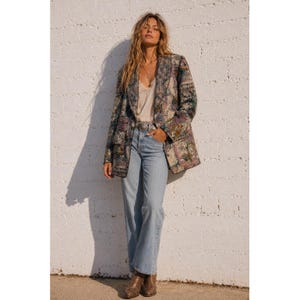 May include: A long-sleeved blazer featuring a multi-colored tapestry pattern with shades of blue, gold, and cream, worn over a light-colored camisole and light-wash blue denim jeans with a silver-toned belt buckle and brown leather boots.