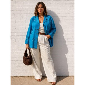 May include: A woman wearing a turquoise blue button-down shirt with floral embroidery, paired with white wide-leg pants and a brown belt. She carries a dark brown woven handbag. The outfit is completed with sandals.
