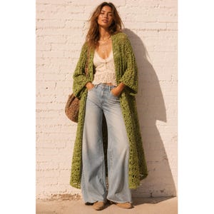 Può includere: Un lungo cardigan all'uncinetto color verde oliva aperto sul davanti con maniche larghe, indossato sopra una canotta in pizzo color crema e jeans a gamba larga in denim chiaro. Una borsa a tracolla intrecciata marrone completa l'outfit.