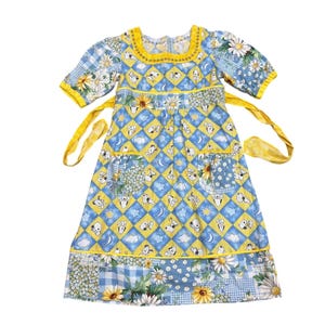 Puede incluir: Vestido de estilo vintage con diseño patchwork en azul, amarillo y blanco, con estampados florales y de personajes de dibujos animados. El vestido tiene mangas cortas, cuello cuadrado y cintas amarillas para atar. La parte inferior del vestido tiene un estampado floral y de cuadros.