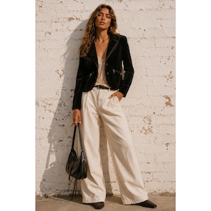 May include: A black velvet blazer with long sleeves and two front pockets, worn over a beige lace-trimmed camisole. The outfit includes high-waisted, wide-leg cream trousers with a black belt and a black leather shoulder bag with a thin strap.