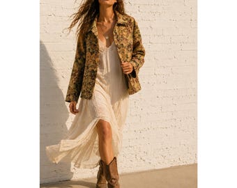 May include: A long-sleeved jacket featuring a multi-colored floral tapestry pattern worn over a cream-colored sleeveless maxi dress with lace detailing. The outfit is paired with brown leather mid-calf boots with a block heel and a silver necklace.