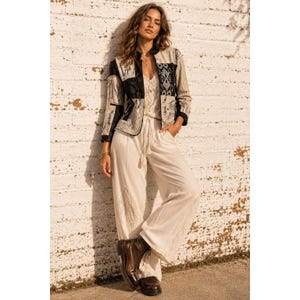 May include: A long-sleeved jacket featuring a black and beige patchwork pattern with a mandarin collar, worn over a cream-colored lace camisole and wide-leg cream trousers with a drawstring waist, paired with dark brown leather lace-up boots.