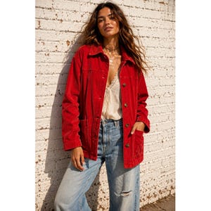 May include: A bright red corduroy jacket featuring a collared neckline, long sleeves, and a button-down front closure. The jacket includes two large patch pockets on the lower front and is worn over a cream-colored top with blue denim jeans.