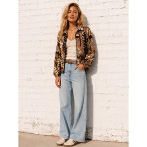 May include: A long-sleeve jacket featuring a brown and black animal print pattern, worn over a cream-colored tank top. The outfit includes light-wash blue denim jeans with a brown leather belt and white sneakers with brown accents.