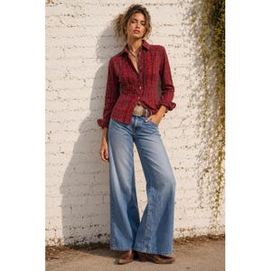 Könnte beinhalten: Ein rotes kariertes Hemd mit Rüschendetails, kombiniert mit hellblauen Jeans mit weitem Bein. Das Outfit umfasst einen braunen Gürtel mit dekorativer Schnalle und braune Stiefel. Das Model steht vor einer weißen Backsteinmauer.