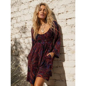 May include: A long-sleeved dress featuring a dark base with a vibrant, multi-coloured paisley pattern in shades of red, blue, and orange. The garment has a gathered, elasticised bodice, a scoop neckline, and a relaxed skirt with side pockets.