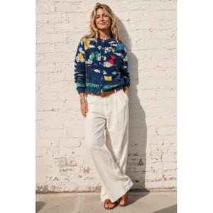 May include: A long-sleeve navy blue knit cardigan featuring a colourful pattern of white clouds and multi-coloured kites, paired with high-waisted cream-coloured wide-leg trousers, a brown leather belt, and brown leather sandals.