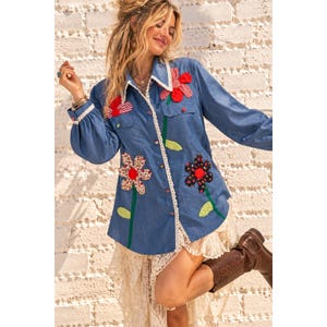 Peut inclure: Une chemise à manches longues en jean bleu avec une bordure en dentelle blanche le long du col et de la patte de boutonnage. La chemise est ornée de grandes applications de fleurs en tissu aux motifs rouges, blancs et noirs avec des tiges vertes.