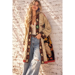 May include: A long, beige open-front duster cardigan featuring a dark trim with a repeating owl pattern. The garment includes a large, detailed owl graphic on the lower side panel, accented by colorful autumn leaves against a black background.