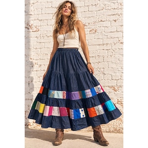 Puede incluir: Una falda larga de mezclilla azul oscuro con un diseño de niveles y una banda horizontal de parches de tela multicolor cerca del dobladillo. La falda tiene una cintura fruncida y una silueta amplia y fluida que llega hasta los tobillos.