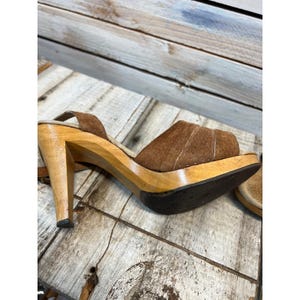 May include: A brown suede sandal with a high wooden heel and platform sole. The upper features a wide strap with subtle perforated detailing. The shoe has a dark brown textured outsole and a light-colored wooden heel and platform base.