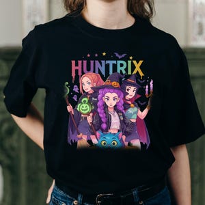 Puede incluir: Camiseta negra con un gráfico colorido de tres brujas de estilo anime, la palabra "HUNTRIX" en letras de arcoíris y un gato azul. Las brujas sostienen objetos mágicos, ideal para Halloween.