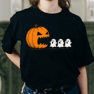Puede incluir: Camiseta negra con un diseño de Halloween. El gráfico muestra una calabaza naranja con una boca dentada, que parece estar comiendo tres pequeños fantasmas blancos. La ilustración tiene un estilo dibujado a mano, ideal para Halloween.