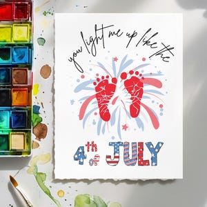 Puede incluir: Una tarjeta blanca con el texto "you light me up like the 4th of July" con huellas rojas y un diseño de fuegos artificiales en rojo, blanco y azul. Pinturas de acuarela y un pincel en el fondo.