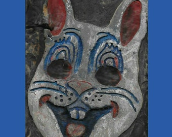 May include: A carved wooden rabbit face with a gray base, blue accents, and red details. The rabbit has large black eyes, a wide smile, and red inner ears. The background is dark and textured.
