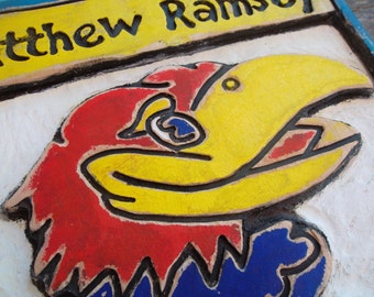 May include: A hand-painted, textured sign featuring a stylized red, yellow, and blue bird emblem. The bird's head is prominently displayed, with the name "Matthew Ramsey" in black lettering on a yellow banner above. The sign has a white background.