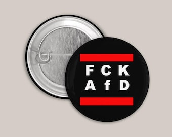 May include: A round pin-back button featuring a black background with two horizontal red stripes and the white text F C K A f D centered between them. The back of the button shows a metallic silver pin mechanism for attachment.