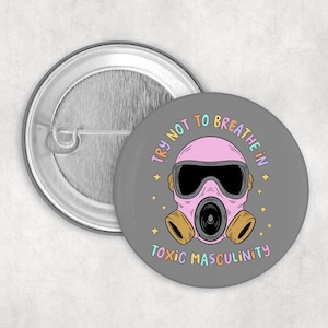 Könnte beinhalten: Ein grauer Button mit einer rosa Gasmasken-Illustration und dem Text "TRY NOT TO BREATHE IN TOXIC MASCULINITY". Die Gasmaske hat schwarze Linsen und beige Filter. Ein zweiter, schlichter Button ist sichtbar.
