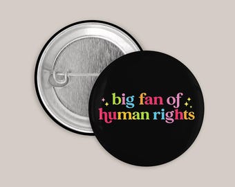 May include: A round pin-back button with a black background featuring the colorful text 'big fan of human rights' in a playful font, accented by small yellow star icons. The back of the button shows a silver metal pin mechanism.