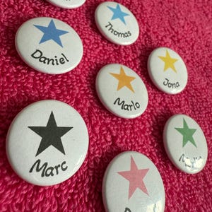 Könnte beinhalten: Eine Sammlung weißer Ansteckbuttons mit bunten Sternmotiven und Namen in schwarzer Schrift. Die Sterne sind in Rot, Blau, Gelb, Orange, Grün und Rosa gehalten. Die Namen sind Sandra, Daniel, Thomas, Marlo, Jona, Marc und Daniel.