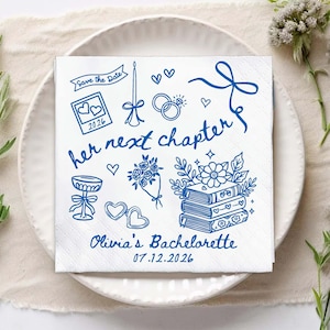 May include: White square napkin with blue illustrations and text. The design includes hearts, rings, flowers, books, and the words "her next chapter" and "Olivia's Bachelorette 07.12.2026". The napkin is on a white plate.