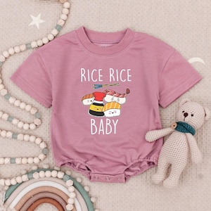 May include: A mauve baby romper with a crew neck and short sleeves. The front features the text "RICE RICE BABY" above a graphic of sushi and soy sauce. The romper is made of soft material and has snap closures.