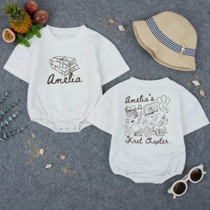 May include: Two white baby rompers with short sleeves. One has the name "Amelia" and a stack of books printed on the front. The other has the text "Amelia's First Chapter" with illustrations on the back. A straw hat and sunglasses are also in the image.