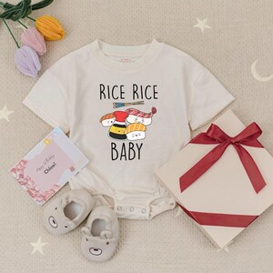 May include: A cream-colored baby onesie with the text "RICE RICE BABY" and a sushi graphic. The image includes a pair of bear-shaped baby shoes, a gift box with a red ribbon, and a birthday card.