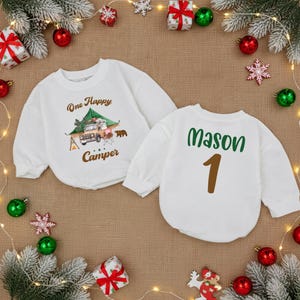May include: Two white baby sweatshirts. One features a camping scene with text that reads "One Happy Camper." The other has the name "Mason" and the number "1" printed on the back. Festive holiday decorations surround the sweatshirts.