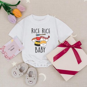 May include: White baby onesie with the text "RICE RICE BABY" and a sushi graphic. The image includes a gift box with a burgundy ribbon, a greeting card, and a pair of gray bear-shaped baby shoes. Floral accents add a decorative touch.