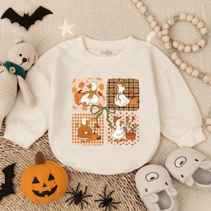 May include: Cream-colored long-sleeved sweatshirt featuring a four-panel design with cartoon geese, pumpkins, and autumn leaves. The design includes a green bow. The brand name "Stofaz" is printed on the upper right. The sweatshirt is surrounded by Halloween-themed decorations.