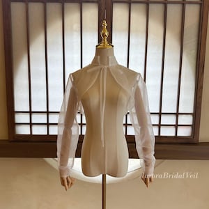 May include: A sheer white bridal bolero with long sleeves and a high neckline, displayed on a mannequin. The bolero has a delicate, airy appearance, with a tie closure at the neck. The background features a window with a grid pattern.