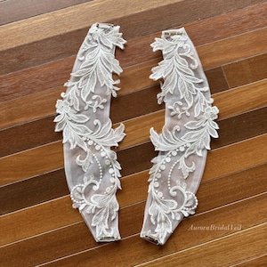 May include: Two white lace bridal sleeve cuffs with floral and leaf patterns. The cuffs are made of delicate lace and feature intricate embroidery with pearl accents. The cuffs are designed to be attached to a wedding dress.