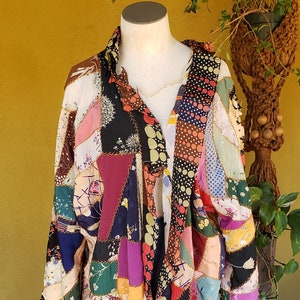 May include: A long, patchwork coat made of various fabrics in shades of pink, red, green, blue, and brown. The coat is sewn together with gold thread and has a frayed hem.