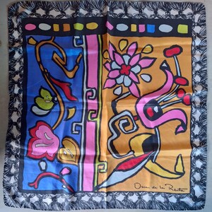 May include: A square silk scarf with a colorful floral design. The scarf features a black border with a white and black pattern, and a tassel fringe. The design includes blue, orange, pink, and yellow floral elements. The designer's signature, Oscar de la Renta, is visible.