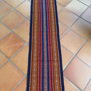 May include: A rectangular, woven textile with a striped pattern. The design features vertical stripes in shades of blue, red, brown, and white. The edges are finished with a dark blue border and fringed ends. This textile could be used as a table runner or decorative accent.