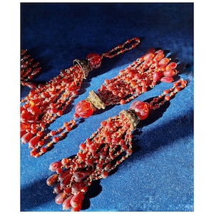 May include: Three red glass tassel ornaments with gold accents. The tassels are made of small, round beads and are attached to a chain.