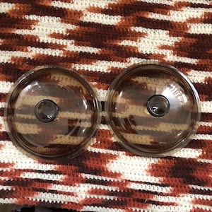 May include: Two clear, round, glass pot lids with dark brown knobs. The lids are transparent, allowing a view of the contents of the pot. The lids are on a brown, white, and orange patterned surface. Kitchenware.