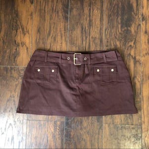 May include: A brown mini skirt with a belt and two front pockets. The skirt has a buckle closure and gold-colored buttons on the pockets. The skirt is made of a solid-colored fabric.