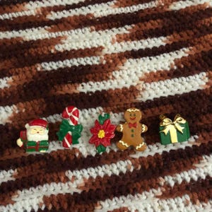 May include: A selection of Christmas-themed brooches. The brooches feature a Santa Claus, a candy cane with a green bow, a poinsettia, a gingerbread man, and a green gift box with a gold bow. They are displayed on a brown, white, and tan crocheted blanket.