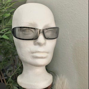 May include: Rectangular sunglasses with dark tinted lenses are displayed on a white mannequin head. The sunglasses have a black frame and are set against a light green background. The image showcases eyewear.