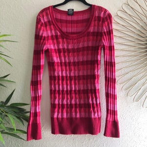 Peut inclure: Un pull en tricot côtelé à manches longues dans les tons de rose et de rouge. Le pull présente une encolure dégagée et un motif rayé. Les manches ont un léger évasement au poignet. Le vêtement est suspendu à un cintre noir.