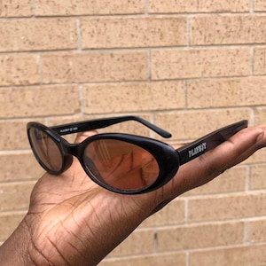 May include: Black oval sunglasses with brown tinted lenses. The sunglasses have the word "PLAYBOY" printed on the arm. The sunglasses are held in a hand.